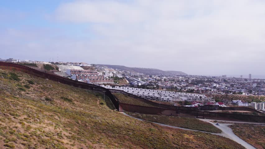 Low-altitude drone view of the U.S.–Mexico border fence cutting through Tijuana’s residential area, contrasting dense city life with border security.
