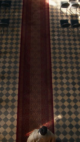 Vertical top view slowmo shot of clergyman wearing majestic golden cope walking confidently red carpet in empty dim lit Catholic church with rows of pews on checkered floor