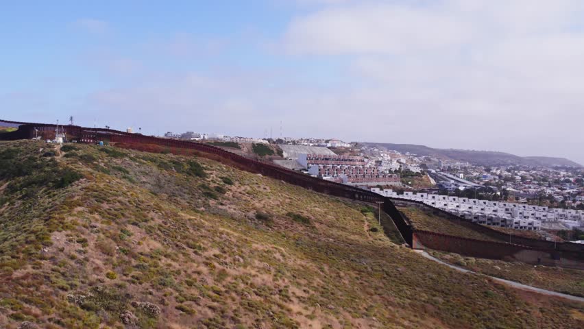 Aerial tracking shot of the U.S.–Mexico border wall as it follows the terrain from desert hills into a dense urban district near Tijuana.