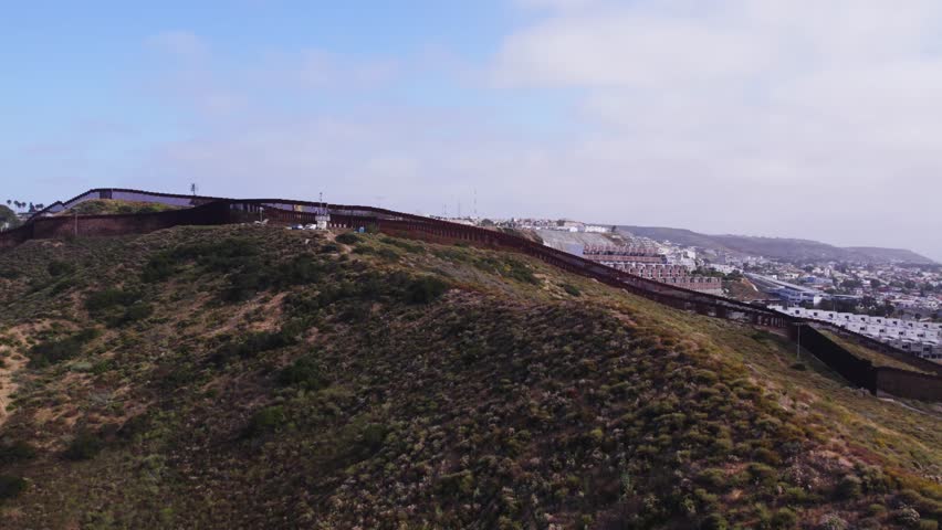 Low-flying aerial tracking shot of the U.S.–Mexico border wall near Tijuana, capturing the contrast between rugged terrain and dense urban development.