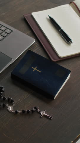 Vertical no people view of wooden table featuring paperbacked bible, prayer beads with cross, notebook and pen, part of laptop