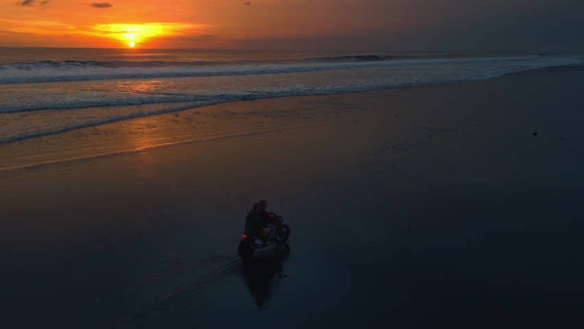 Couple riding a motorcycle on black sand beach at sunset, with a red signal flare, fire. Ocean waves and tropical scenery, romantic vibe. Man, woman driving moto bike on coastline of Bali. Road trip