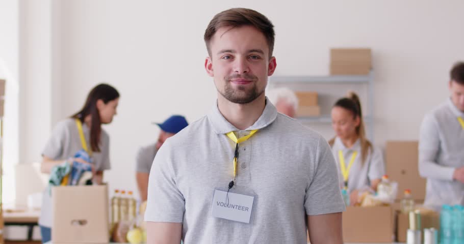 Male volunteer shows thumbs up against background of people sorting and organizing food donations. Young Caucasian man wearing t-shirt and with badge recommends volunteering. 4k footage.
