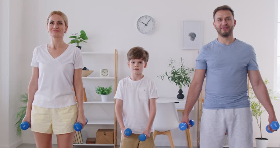 Portrait of sporty happy family with child boy doing sport exercises holding dumbbells and looking cheerful at camera standing at home. Family sport concept. 4k video. Slow motion video
