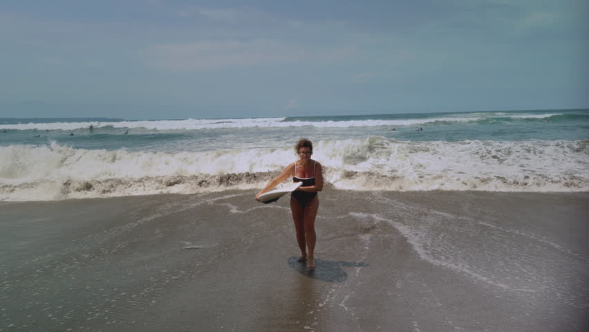 Active mature woman in wetsuit with surfboard at the beach, enjoying a healthy lifestyle. Old senior female doing surfing on Bali. Elderly lady holding surf board. Tropical coast in asia, big wave.