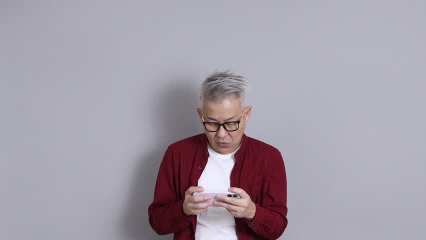 The senior East Asian man, wearing a red shirt, is standing against a grey background.