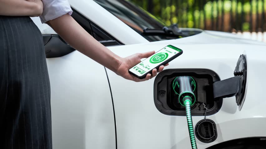 Businesswoman holding smartphone display battery status interface by smart EV mobile application while eco-friendly EV car recharging electricity from charging station in car park. Peruse