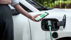 Businesswoman holding smartphone display battery status interface by smart EV mobile application while eco-friendly EV car recharging electricity from charging station in car park. Peruse - Powered by Shutterstock - Get 15% off with code: PIKWIZARD15