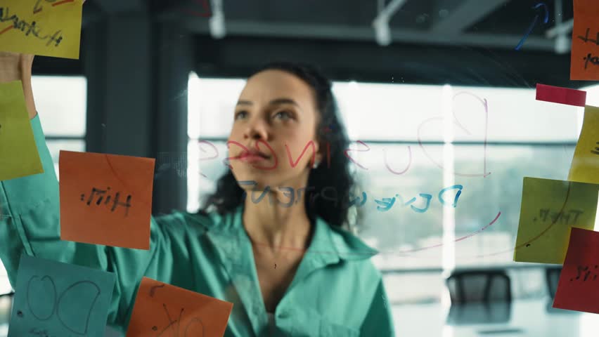 Portrait of smart professional hispanic businesswoman writes marketing ideas on glass board with colorful sticky notes. Young manager drawing mind map to analysis business plan. Closeup. Manipulator.