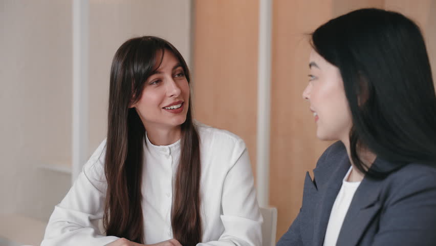 Two smiling women enjoying lighthearted conversation while sitting face to face in relaxed indoor setting. Sharing stories or catching up during lunch break with friendly expressions and warm energy.