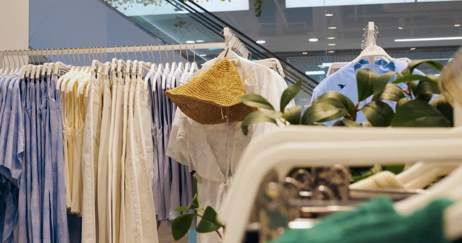 Light summer clothing and straw hat hanging on white hangers inside modern retail space, green plant adding natural accent to fashion display. Concept fashion clothes