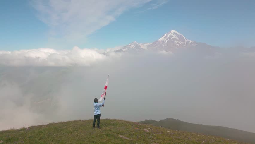 Aerial panning view Georgian woman mountains lover tourist or visitors hold giant georgian country Sakartvelo flag on mountain peak viewpoint outdoors caucasus mountains. Travel tourism georgia banner
