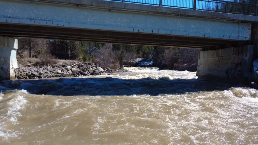 Drone flying under a road bridge where a raging brownish river flows, swollen by the spring flood and the melting snow from the surrounding forest under a blue sky. Tartigou River, Quebec, Canada.