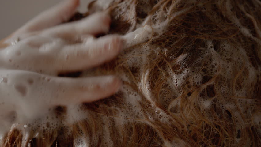 Woman is in a shower, lathering her hair with shampoo and rinsing it out. Water cascades down as she enjoys her cleansing routine amidst the foam and suds.