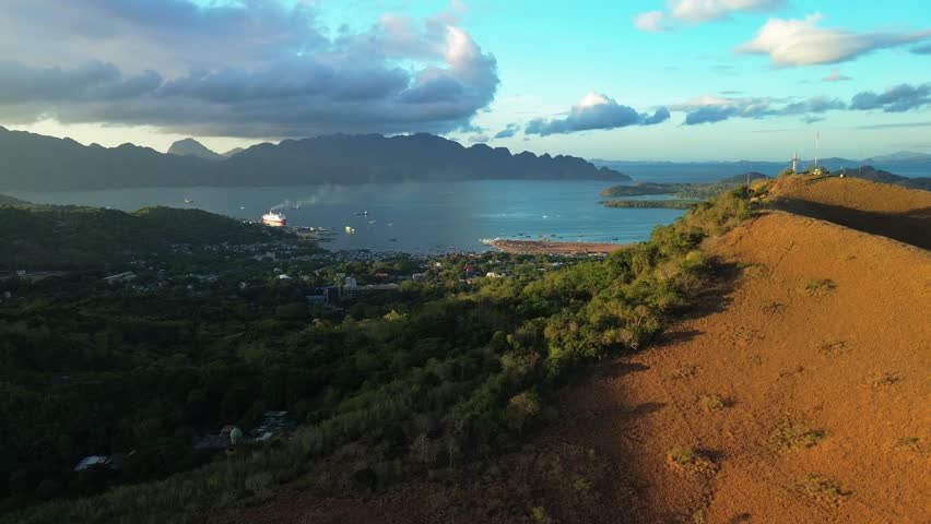 Scenic sunrise shot over Coron town from Mount Tapyas, with a large ferry arriving from Manila as golden light touches the hills and sea.
