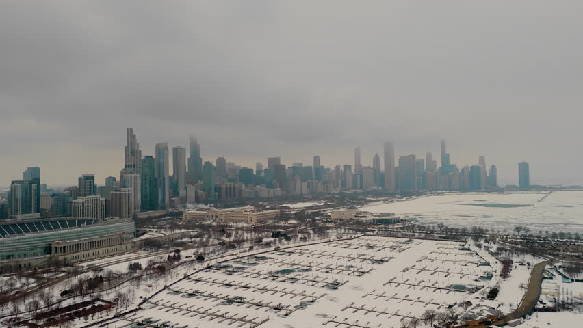 Aerial view of snowy marina on urban Chicago skyline. Frozen harbor, towering buildings in the background. Empty docks with patches of icy green water through the snow