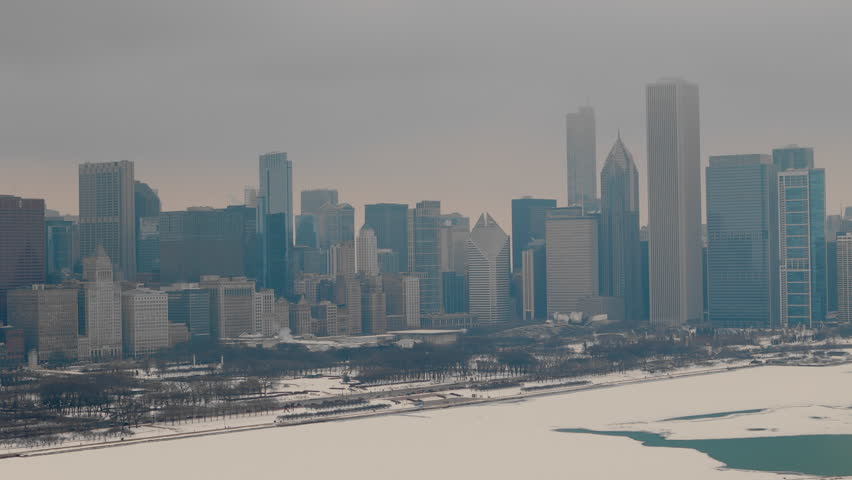 Th aerial view of a snowy urban Chicago skyline. Frozen harbor, towering buildings in the background. Patches of icy green water through the snow