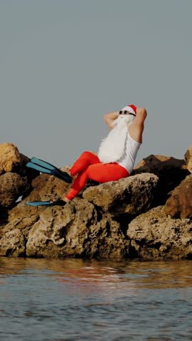 Santa Claus sunbathes. Santa Claus having fun. Funny Santa, in sunglasses and flippers sitting on rocky beach by the sea. Santa Claus summer vacation