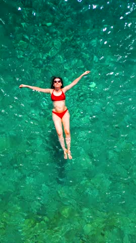Woman in red swimsuit lays on her back in the clear sea, top down view