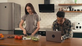 Multicultural couple enjoying a calm morning in the kitchen - young woman slicing vegetables for breakfast while her partner works on a laptop. High quality 4k footage - Powered by Shutterstock - Get 15% off with code: PIKWIZARD15