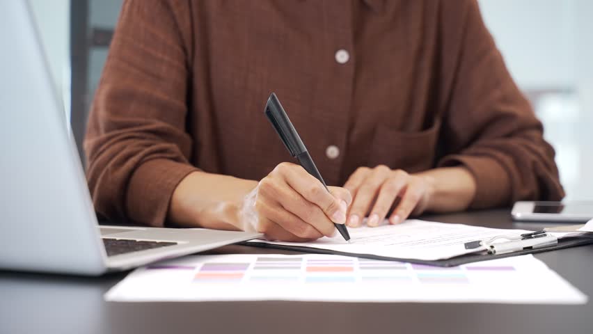 Close up of a female hand filling out documents with a pen using a laptop while sitting at desk at workplace in a business office. Businesswoman is engaged in paper work, making financial calculations
