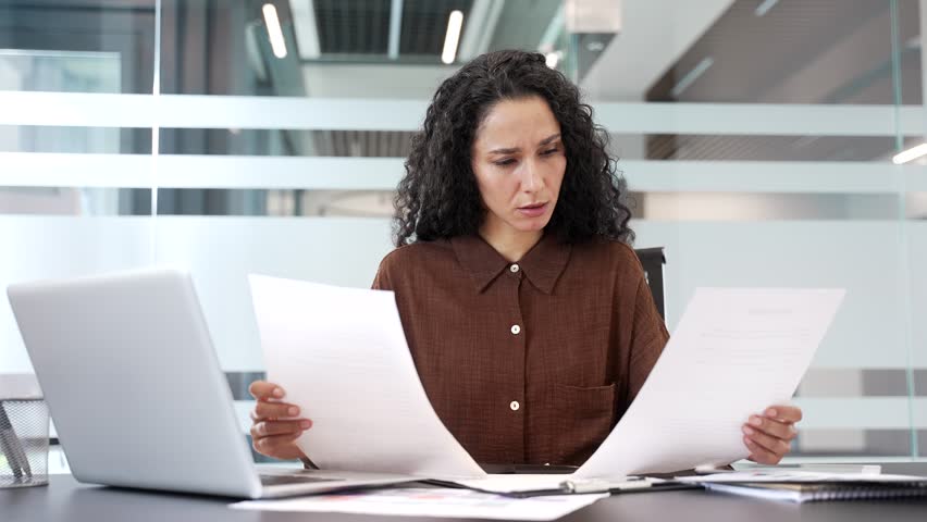 Confused businesswoman having difficulty with paperwork sitting at workplace in business office. Puzzled female employee looks at the documents and cannot understand the problem and looking at camera