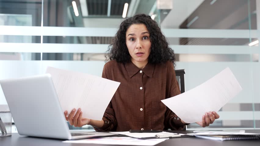 Confused businesswoman having difficulty with paperwork sitting at workplace in business office. Puzzled female employee looks at the documents and cannot understand the problem and looking at camera