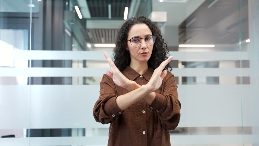 Serious businesswoman showing crossed arms, X sign, shaking head NO while standing in a modern business office looking at camera. The woman demonstrates a gesture of prohibition, denial, disagreement