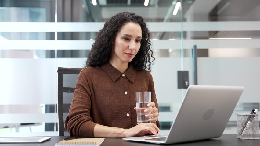 Young businesswoman drinks water from a glass while sitting at desk at workplace in modern business office. Joyful smiling female employee feeling relieved, enjoying clean cool drink, relaxing resting