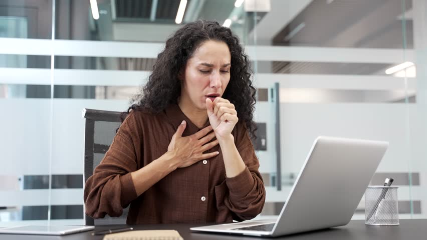 Sick businesswoman coughing while sitting at a desk at a workplace in a modern business office. Unhealthy young female employee is unwell. She has the first signs of a cold, virus, bronchitis or flu