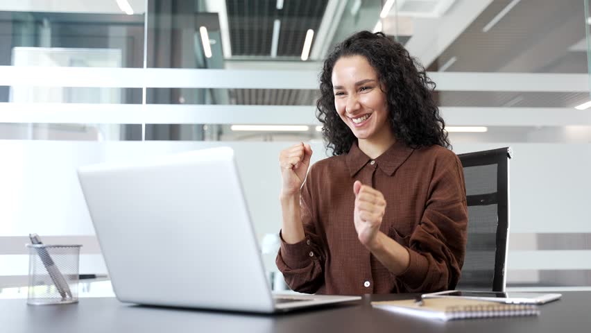 Satisfied happy businesswoman reading good news on laptop sitting at workplace in business office. Smiling female employee rejoices at a pleasant notification, glad at positive message on computer