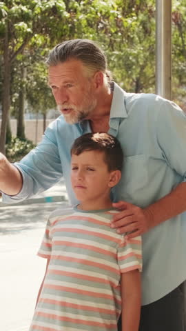 Vertical video, Grandfather and grandson wait at a bus stop. Grandpa has his arm around his grandson and points something out across the street. The boy looks in that direction.