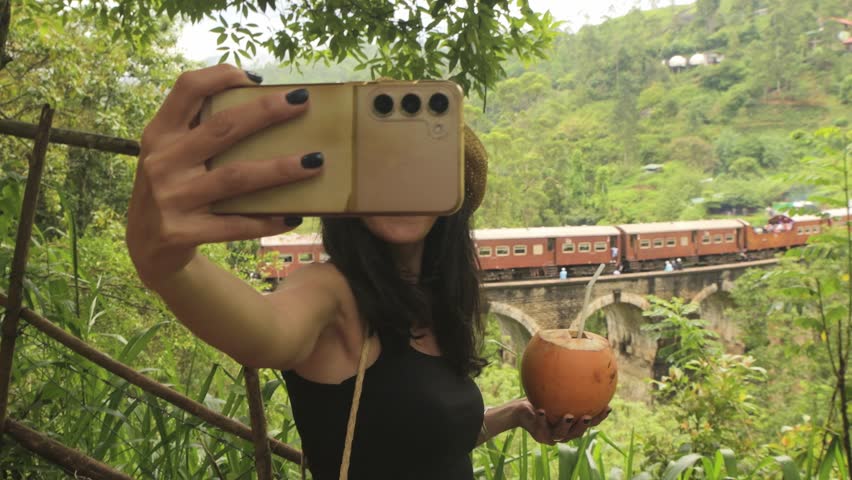 A young woman enjoys a tropical coconut while posing at a scenic viewpoint overlooking the iconic Nine Arch Bridge near Ella, deep in the lush highlands of Uva Province, Sri Lanka