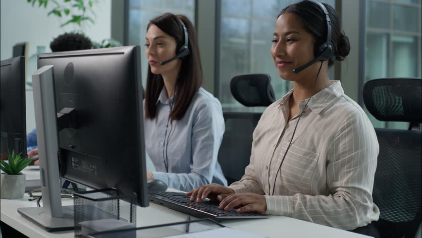 Three business people multiethnic Indian Caucasian women African man headset call center service operators work indoor callcenter talk client using pc computer writing notes helpful customer service