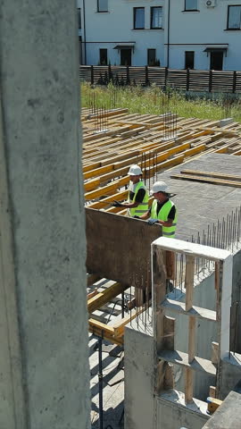 Aerial View of Construction Workers at Building Site. Aerial view captures two construction workers in high-visibility vests as they maneuver a large wooden board across a building site.