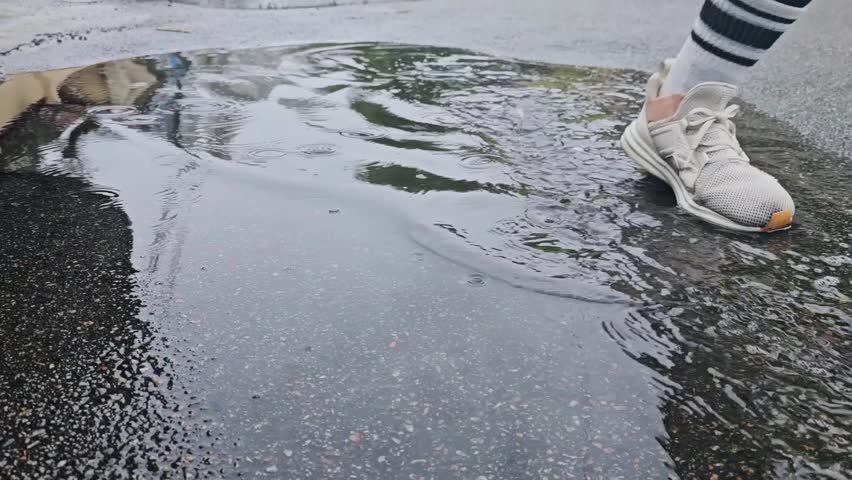 Close up slow motion shot of legs in white sneakers stepping into muddy puddle. Sad woman walking in rain, making splash.
