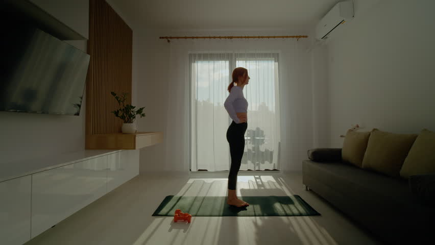 A woman practices stretching and pilates on a mat in her living room filled with sunlight. She embraces a healthy lifestyle with exercises using dumbbells during the morning.