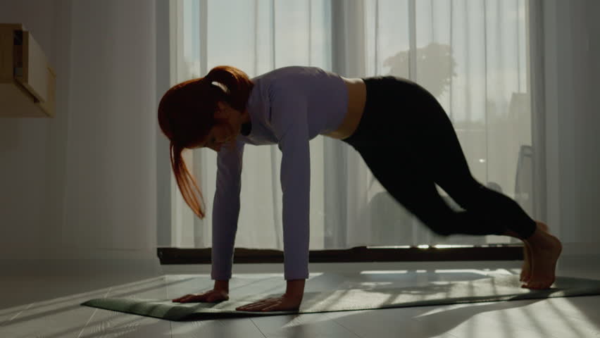 A woman practices yoga and pilates on a mat in her bright living room. Stretching and exercising in the morning sunlight, she embraces a healthy lifestyle.