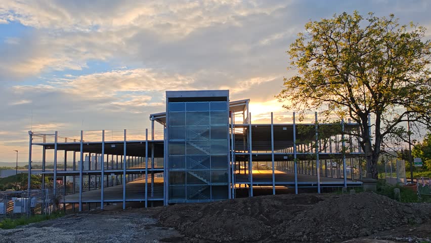 Multi-level parking garage under construction beside factory at city edge, steel frame and concrete in progress.