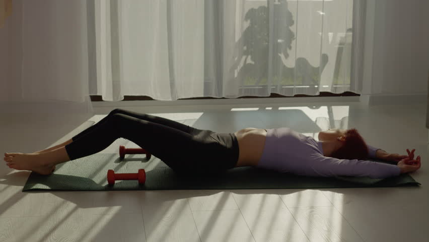 A woman with red hair lies on an exercise mat in her living room, taking a well-deserved break after completing her morning workout with dumbbells and stretching.
