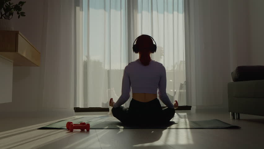 A woman enjoys a peaceful morning meditation while wearing headphones in her sunlit living room. She sits cross-legged, creating a serene atmosphere for relaxation.