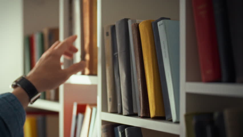 A hand is reaching for a book on a shelf filled with various colored volumes. This action takes place in a warm, inviting library setting, perfect for reading and exploration