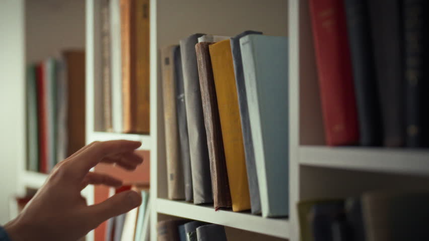 A person is carefully picking a book from a neatly arranged shelf in a quiet library. The setting suggests a peaceful environment perfect for reading and studying