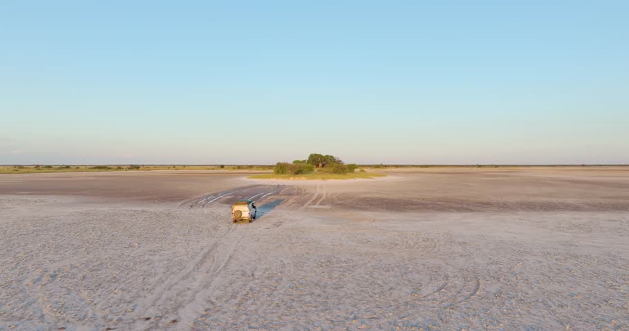 Drone footage of 4x4 off-road vehicle crossing salt pan toward tree-covered island in Nxai Pan, Botswana. Ideal for: travel content, safari promotion, overlanding, remote Africa landscapes.  