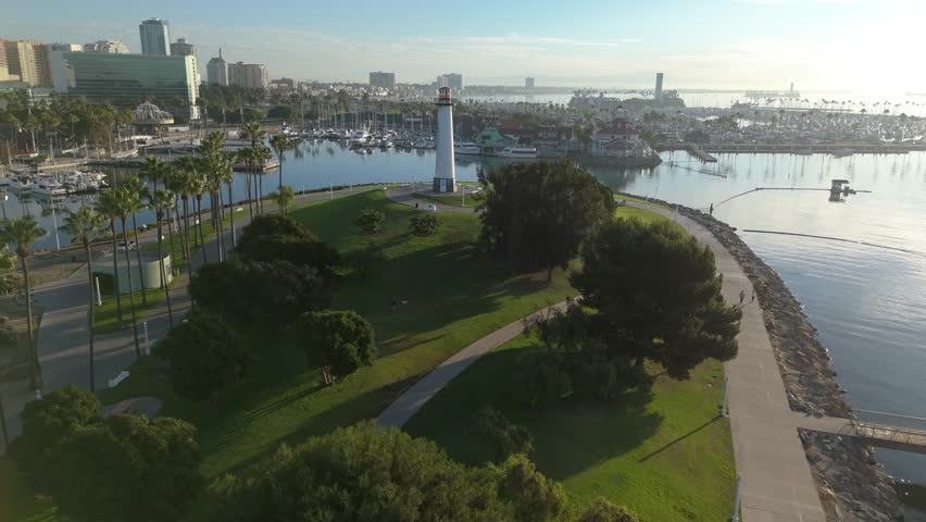 Aerial establishing shot of Lions Lighthouse at Shoreline Park in Long Beach, California, as dawn lights up the peaceful harbor and stunning skyline.