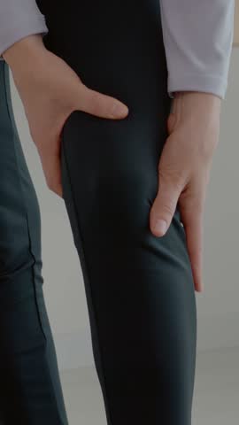Vertical Screen: A woman holds her knee in discomfort while sitting on the floor of her living room. She seems to be dealing with leg pain during her fitness routine.