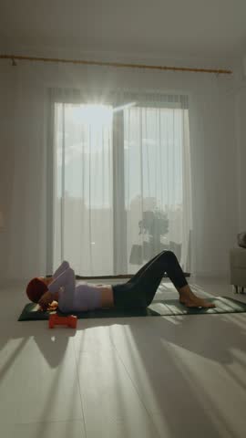 Vertical Screen: A woman practices abdominal exercises on a mat in her sunny living room. The fresh morning light enhances her focused workout session with dumbbells and calm energy.