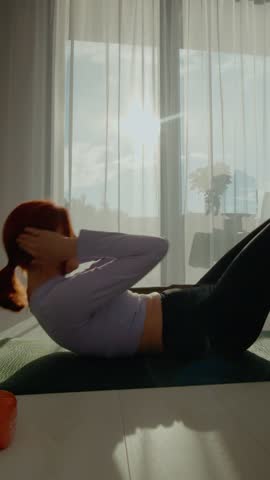 Vertical Screen: A woman practices abdominal exercises on a mat in her sunny living room. The fresh morning light enhances her focused workout session with dumbbells and calm energy.