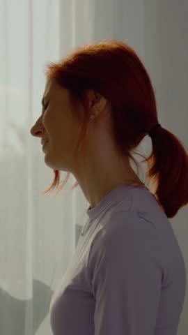 Vertical Screen: A redhead woman sits comfortably in her living room, gently massaging her neck to soothe discomfort from muscle pain caused by strain or dislocation.