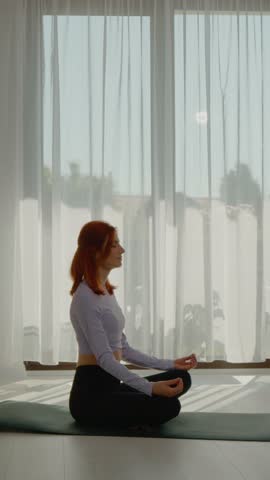 Vertical Screen: A redhead woman engages in yoga and meditation in her living room as the sun rises, focusing on her breath and mental balance against a serene window backdrop.
